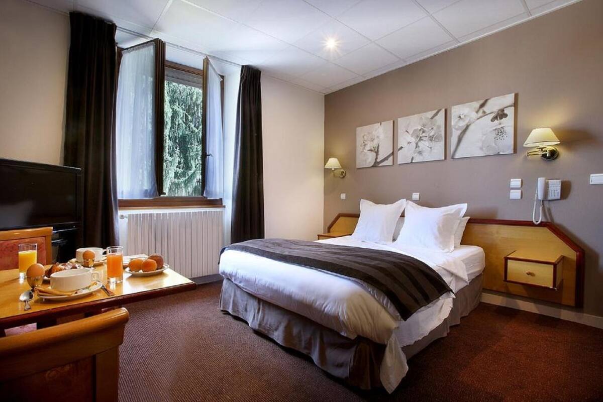 Een hotelkamer met een bed, nachtkastje, lamp, kunst aan de muur en een raam met gordijnen.