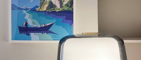 Ein Zimmer mit einem gerahmten Poster einer malerischen Flusslandschaft, einer weißen Lampe und einem Bücherregal mit Büchern.