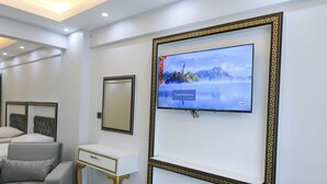 A modern hotel room with a large flat-screen TV mounted on the wall, a grey sofa, a white bedside table, a gold-framed mirror, and a small bench. - Istanbul Hotel & Guesthouse  (Istanbul)