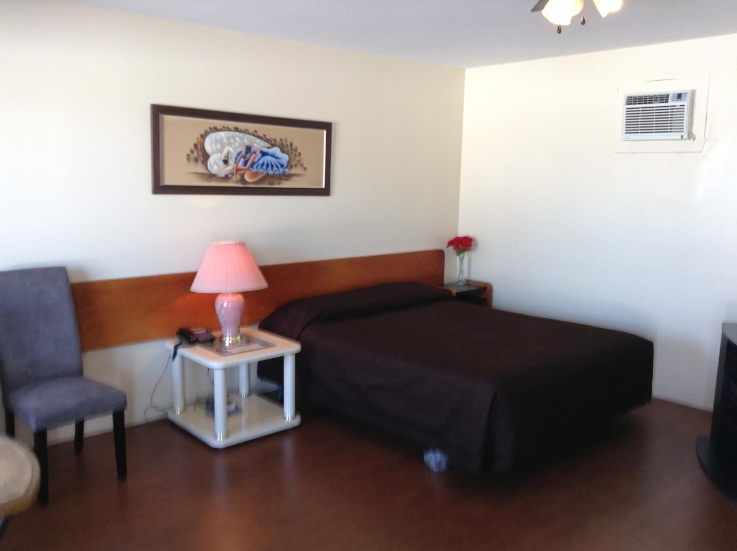 A hotel room with a bed, a chair, a bedside table with a lamp, a framed picture on the wall, and an air conditioning unit.