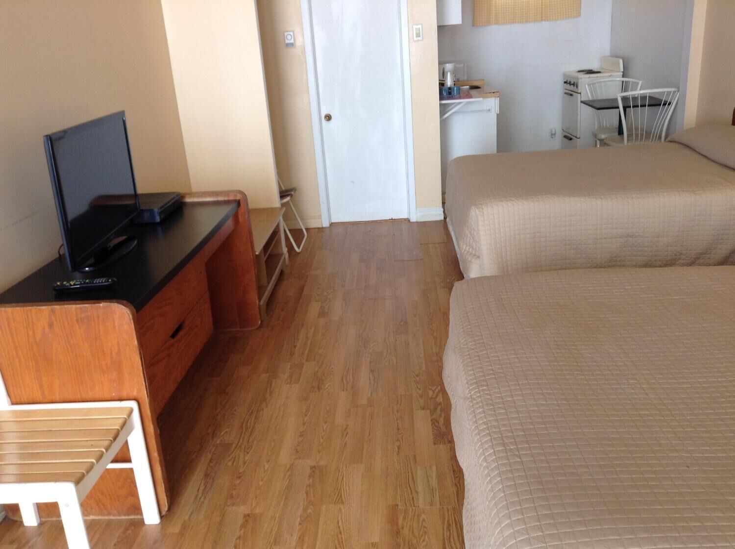A hotel room with a wooden desk, a television, a bed, a small kitchenette, and a white chair.