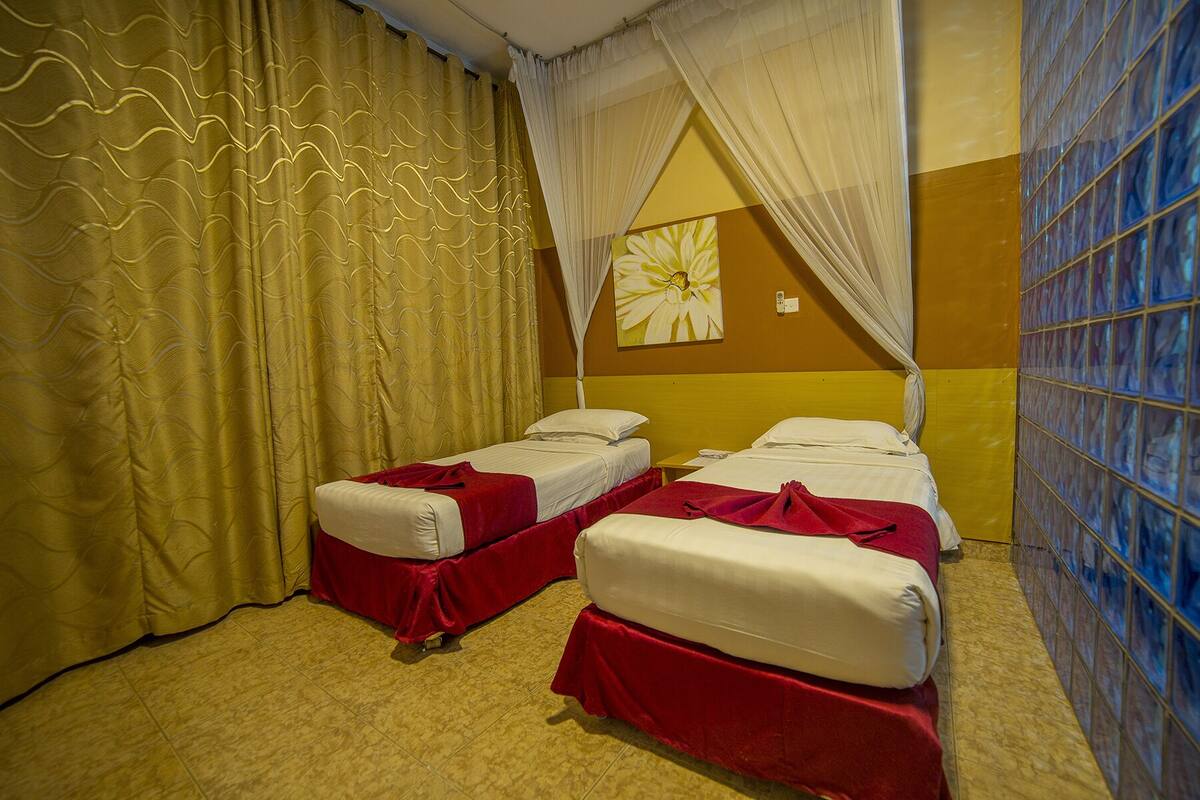 Habitación de hotel con dos camas individuales, un ventanal grande con vidrio azulado, pared amarilla y naranja, y un cuadro grande de flores.