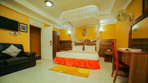 A bedroom with a four-poster bed, a desk, a chair, a television, and a fan. - Eureka Place Hotel (Kampala)