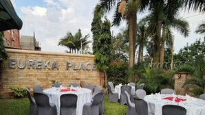 An outdoor dining area with tables and chairs, a sign reading "EUREKA PLACE," and palm trees in the background. - Eureka Place Hotel (Kampala)