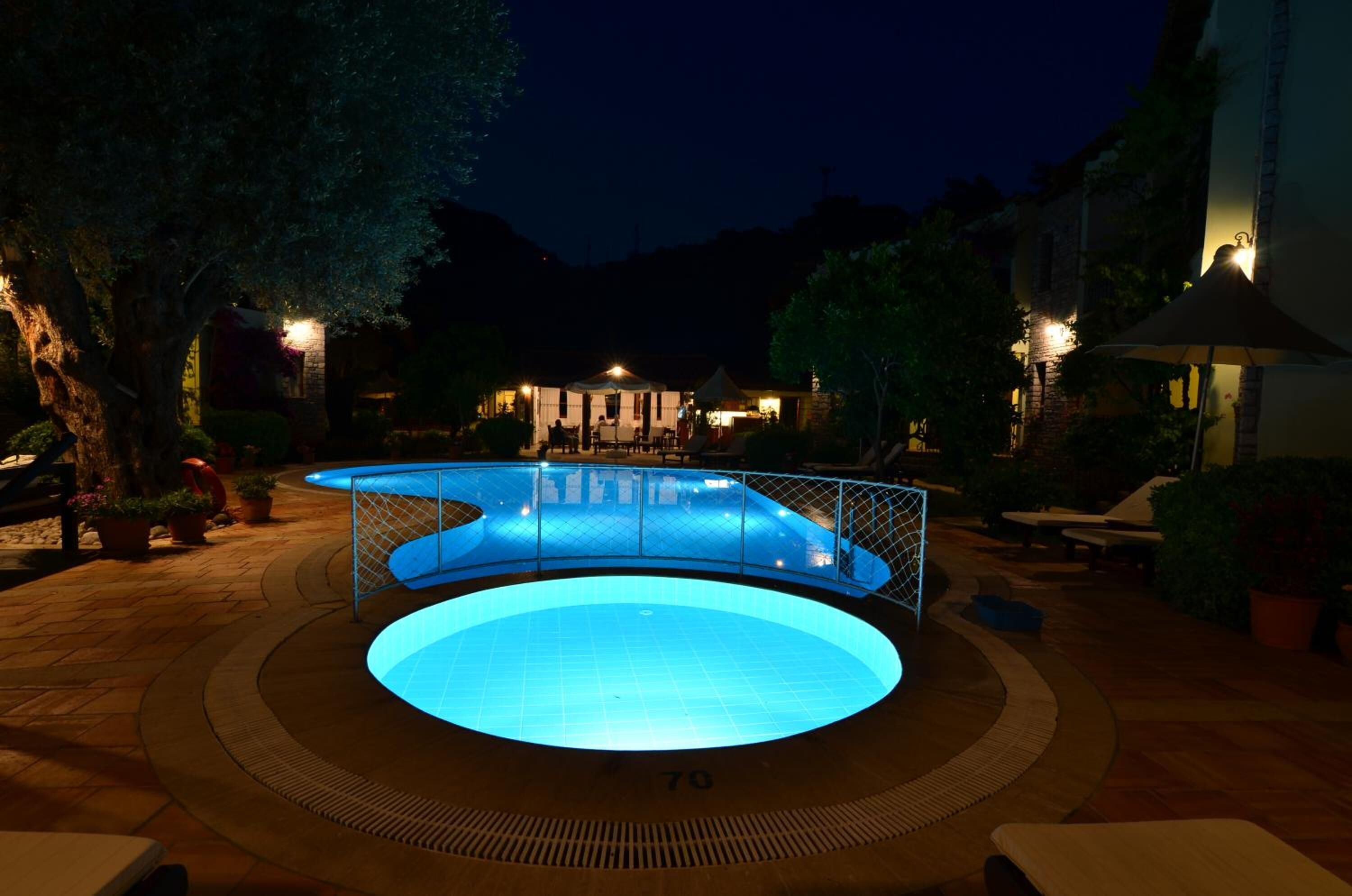 Una piscina con diseño circular, rodeada de sillones de descanso y plantas en macetas, con un edificio al fondo.