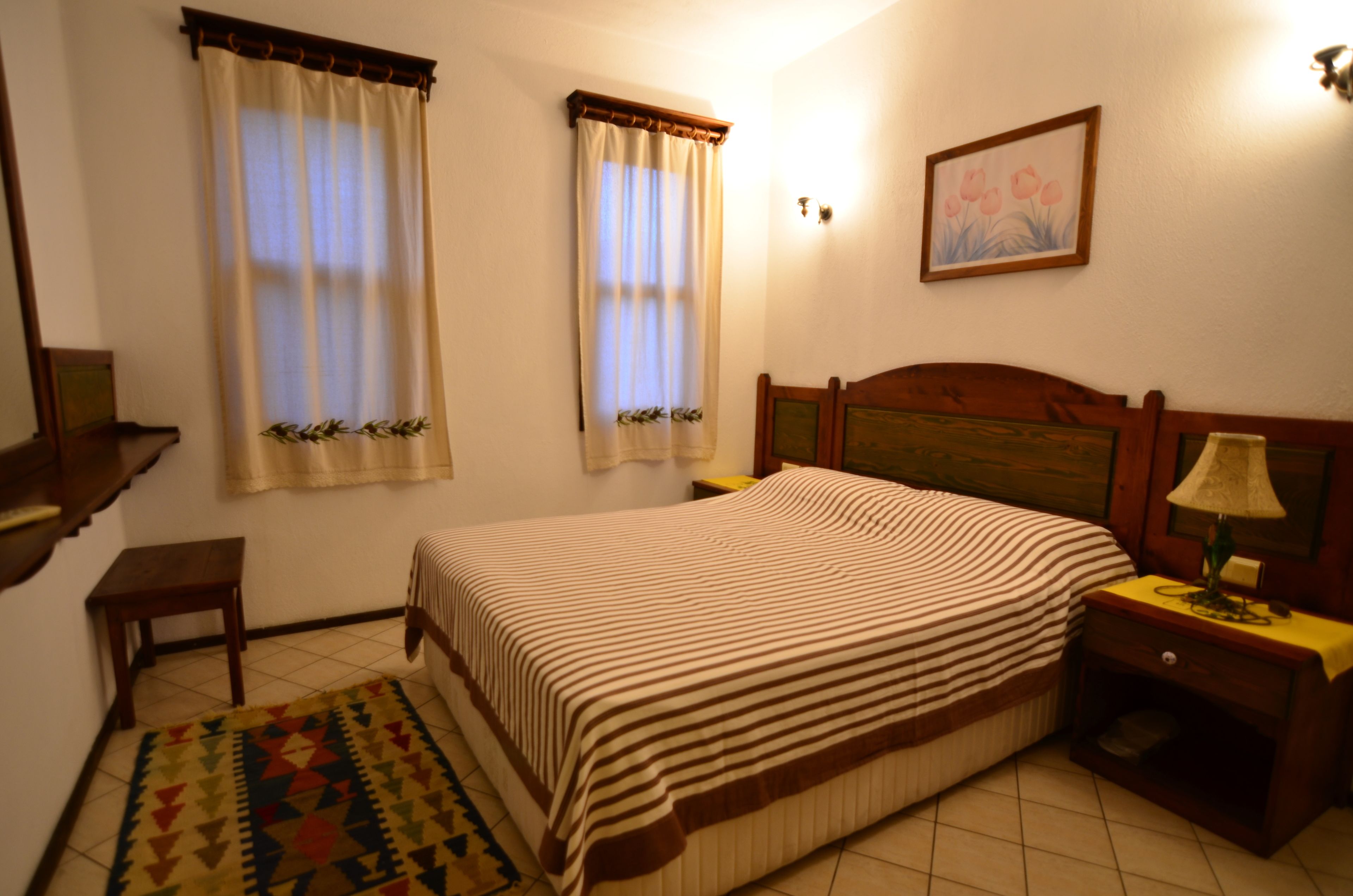 A bedroom with a bed, a nightstand, a lamp, a rug, two windows with curtains, and a wall-mounted picture.