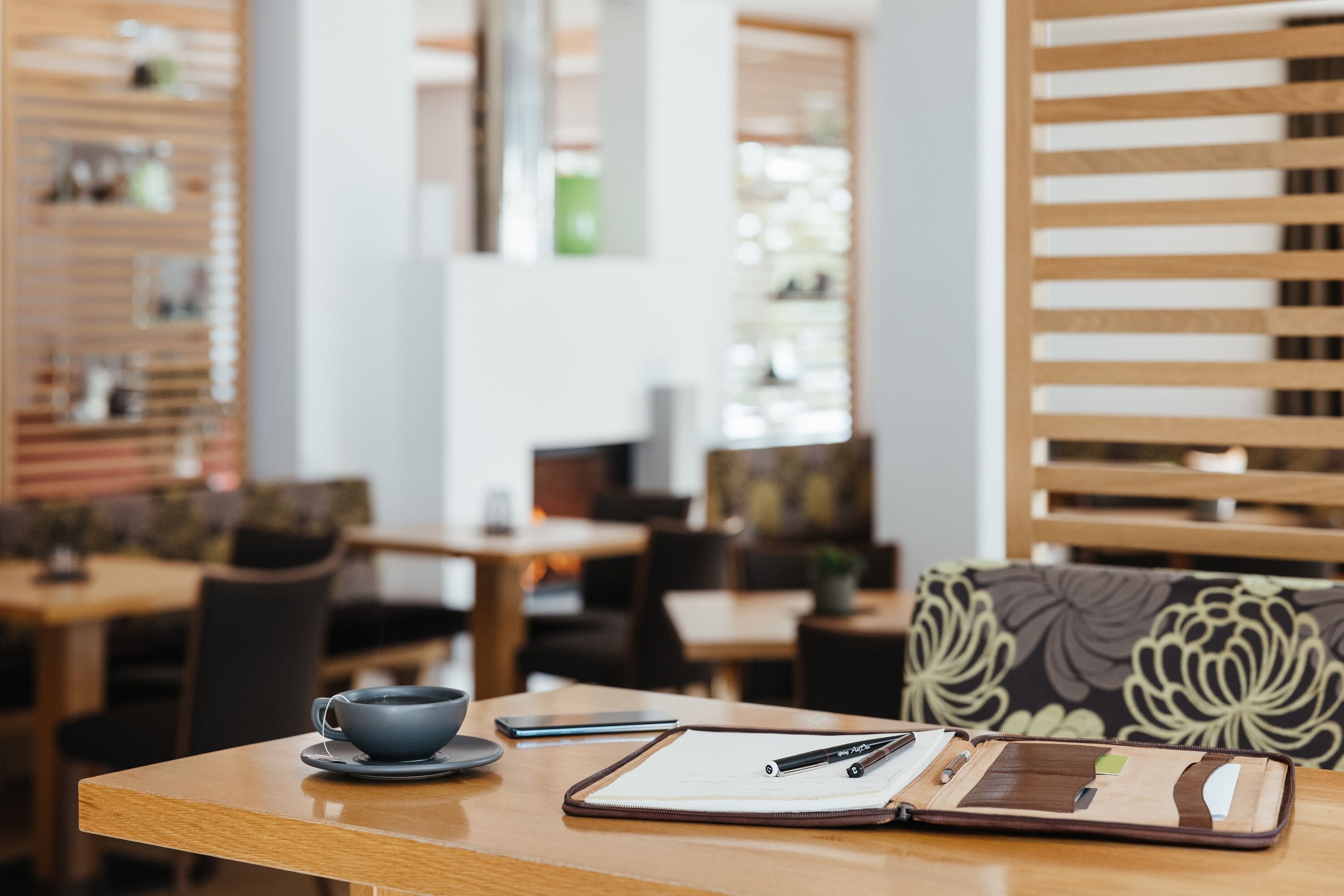A wooden table with a notebook, pen, and cup in a cafe setting.