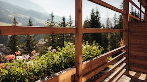 Un balcon en bois, doté d’une balustrade en bois, surplombe un jardin où s’épanouissent différentes espèces de fleurs, avec un paysage montagneux en toile de fond.