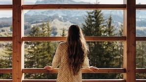 Une personne se tient près d’une grande fenêtre, d’où l’on peut admirer un paysage de montagnes et d’arbres.