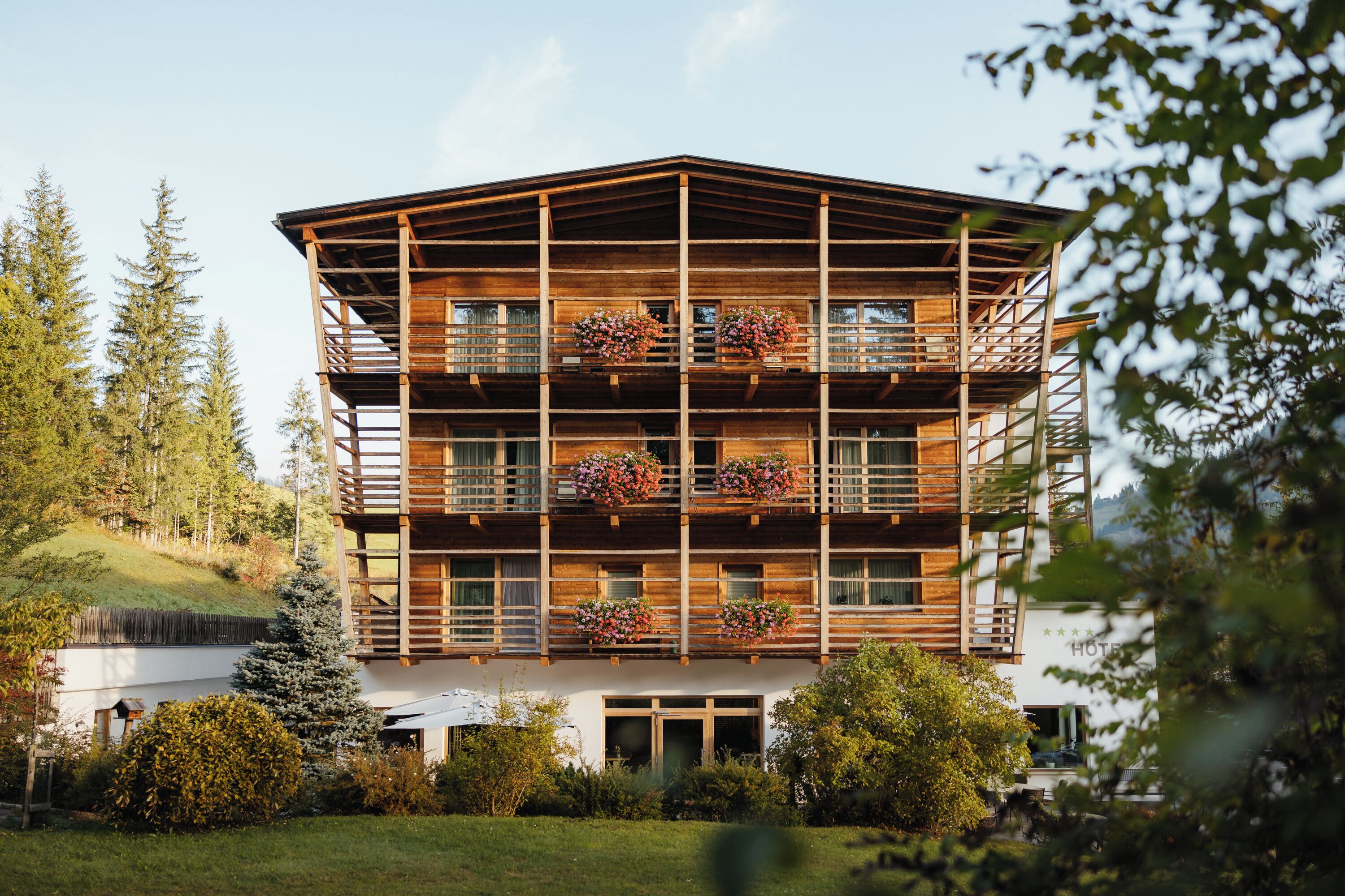 A multi-story wooden building with balconies, surrounded by trees and a grassy area.