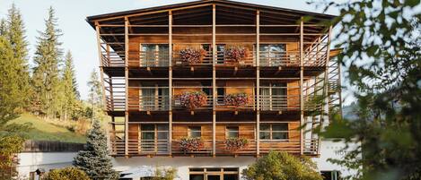 A multi-story wooden building with balconies, surrounded by trees and a grassy area.