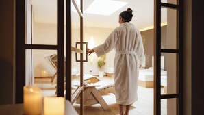 Une personne vêtue d’un peignoir blanc se promène dans une pièce dont le sol est en bois, où se trouvent une table de massage et des bougies.