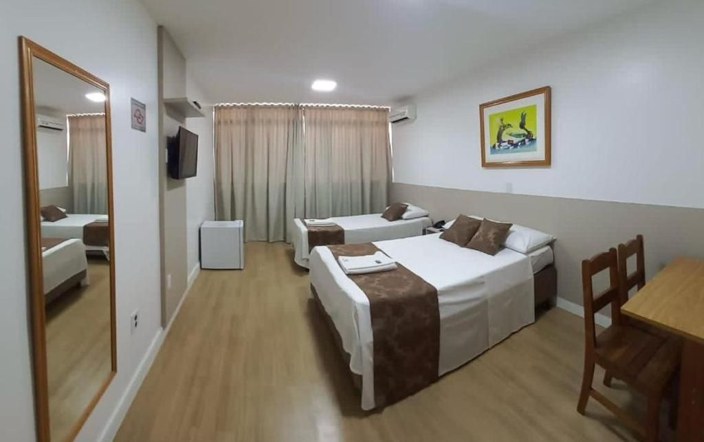 Quarto de hotel com duas camas de solteiro, uma mesa de jantar de madeira, uma cadeira, televisão e um quadro na parede.