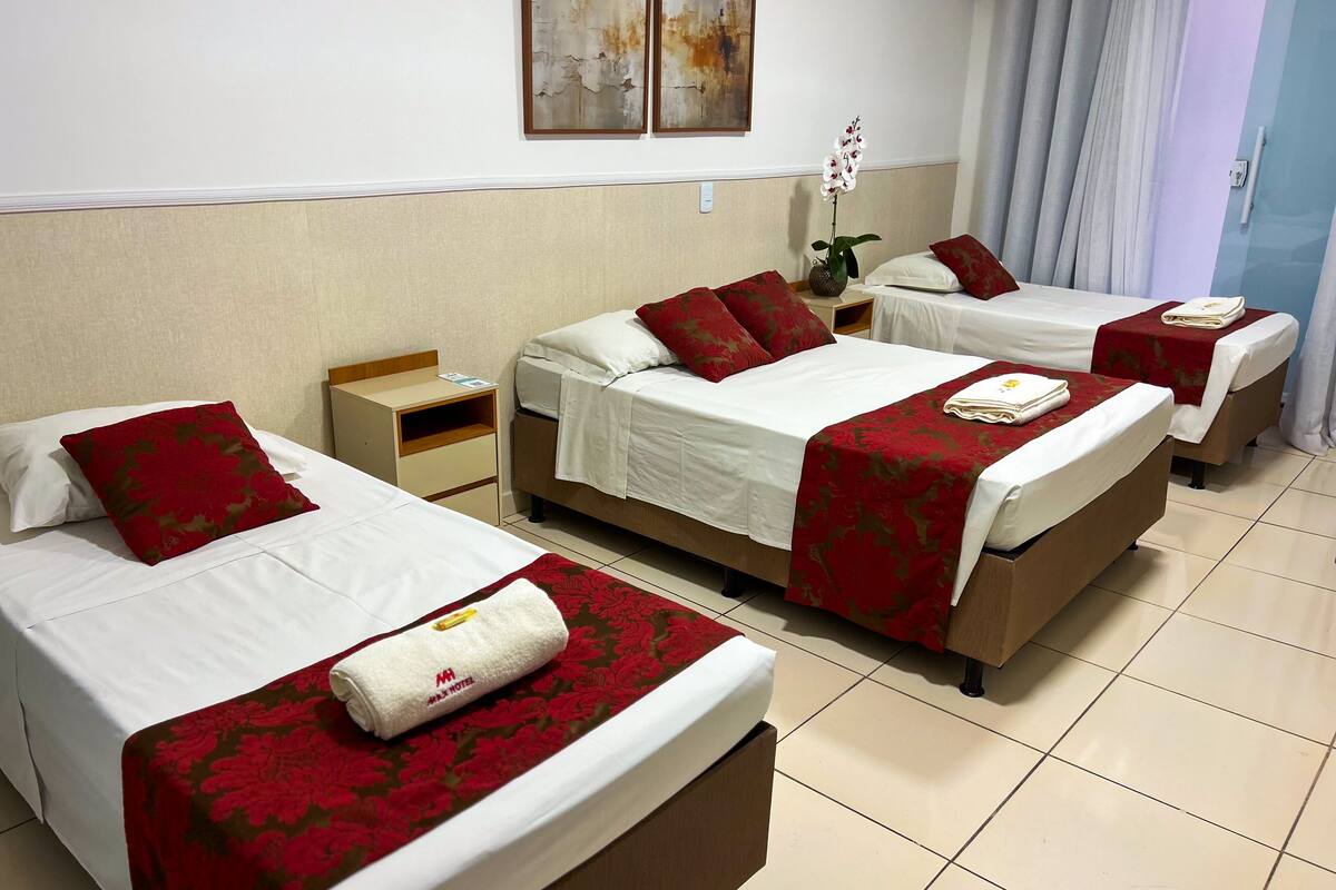 Habitación de hotel con tres camas, cada una con sábanas blancas y cubrecamas con estampado floral rojo. Hay una mesita de noche de madera con una lámpara y un jarrón con flores.