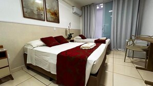 A hotel room with two beds, a desk, and a chair. There is a window with curtains, a wall-mounted air conditioner, and a small table with a vase of flowers. - Max Hotel (Nucleo Bandeirante)