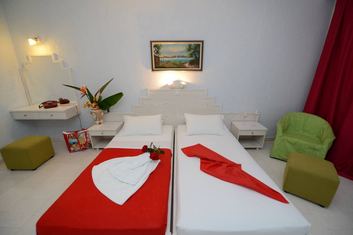 A double room with two beds, a nightstand, a mirror, a small table with a vase of flowers, a framed picture on the wall, and a red curtain.