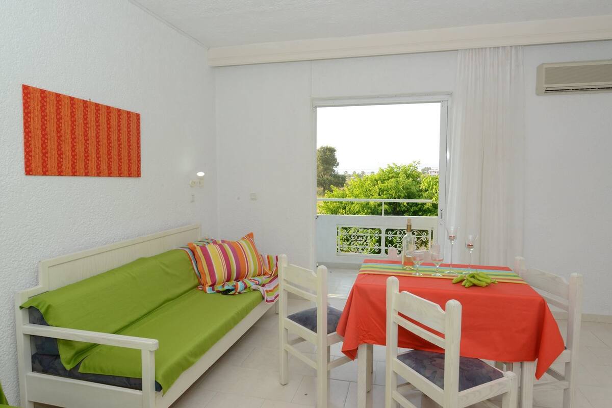 A room with a bed, a dining table with chairs, a window with a view of greenery, and a wall-mounted air conditioning unit.