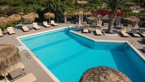 Zona de piscina con sillones de descanso y sombrillas de paja, rodeada de vegetación y vistas a edificios lejanos.