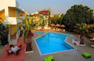 Piscina con sillones de descanso y sombrillas, rodeada de palmeras y un edificio con balcones.