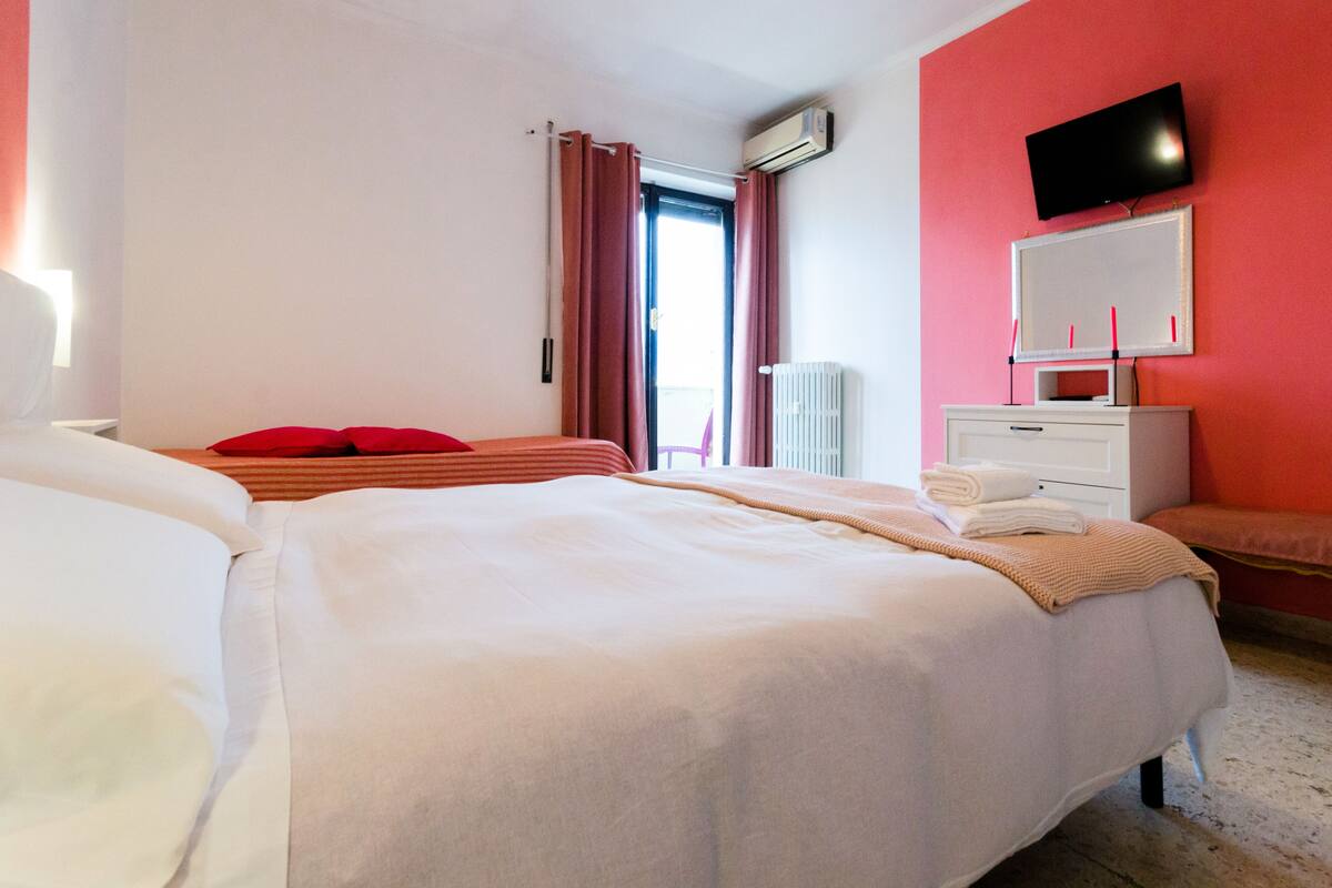 Habitación de hotel con una cama grande, una mesita de noche, un televisor montado en la pared y un ventanal con cortinas.