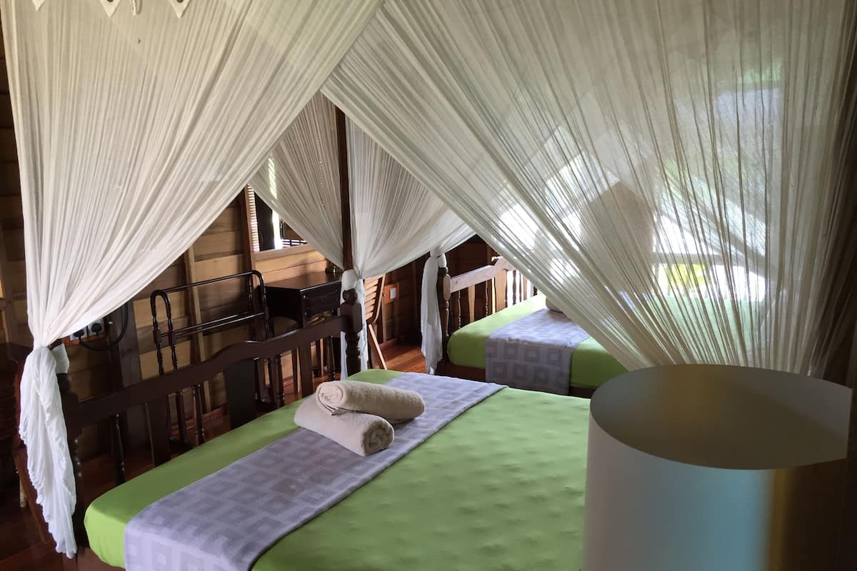 A room with two beds, each with a green bedspread and a white pillow. There is a wooden chair and a wooden bench. The room has a wooden floor and a wooden railing.