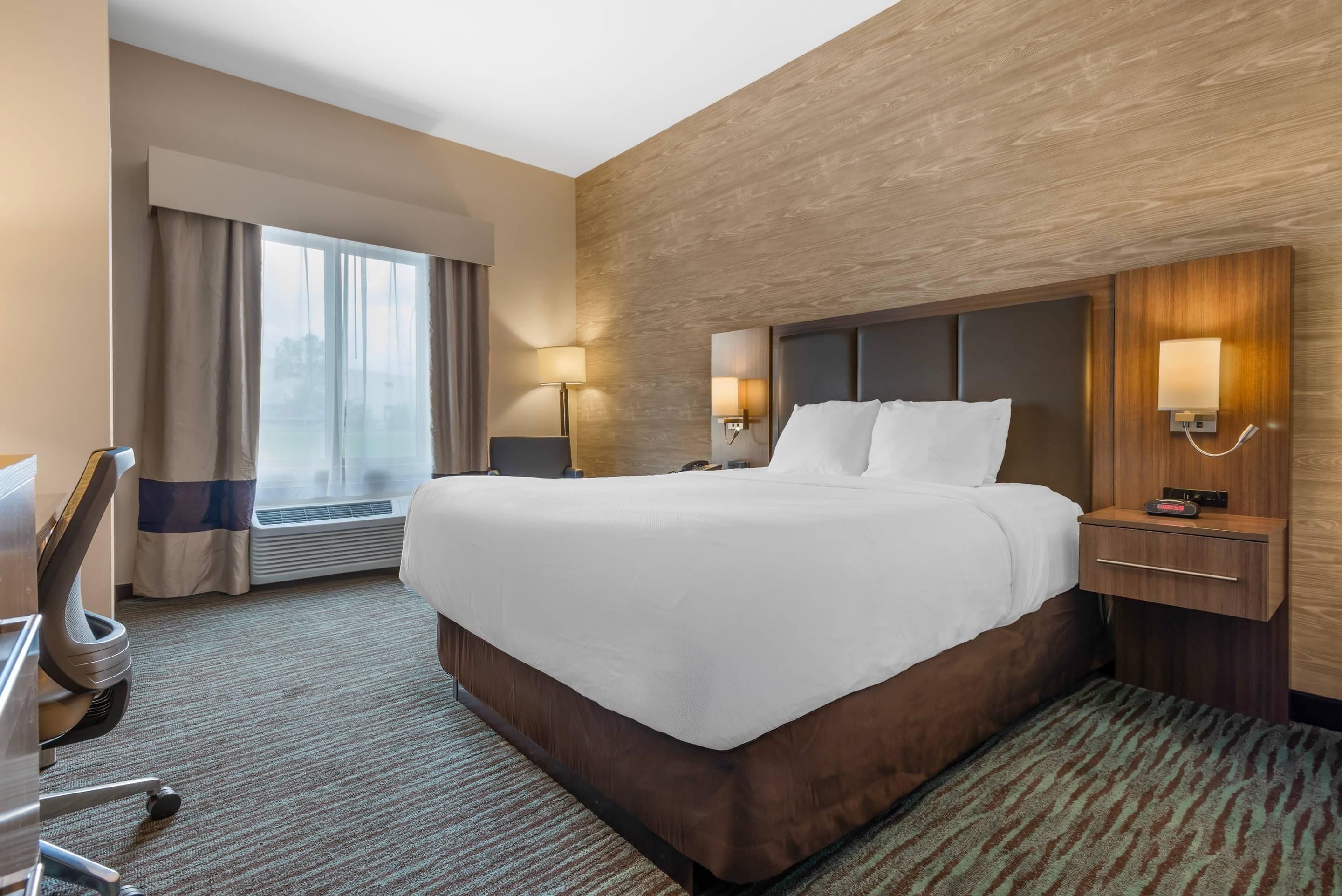 A hotel room with a large bed, two bedside lamps, a desk with a chair, and a window with curtains.