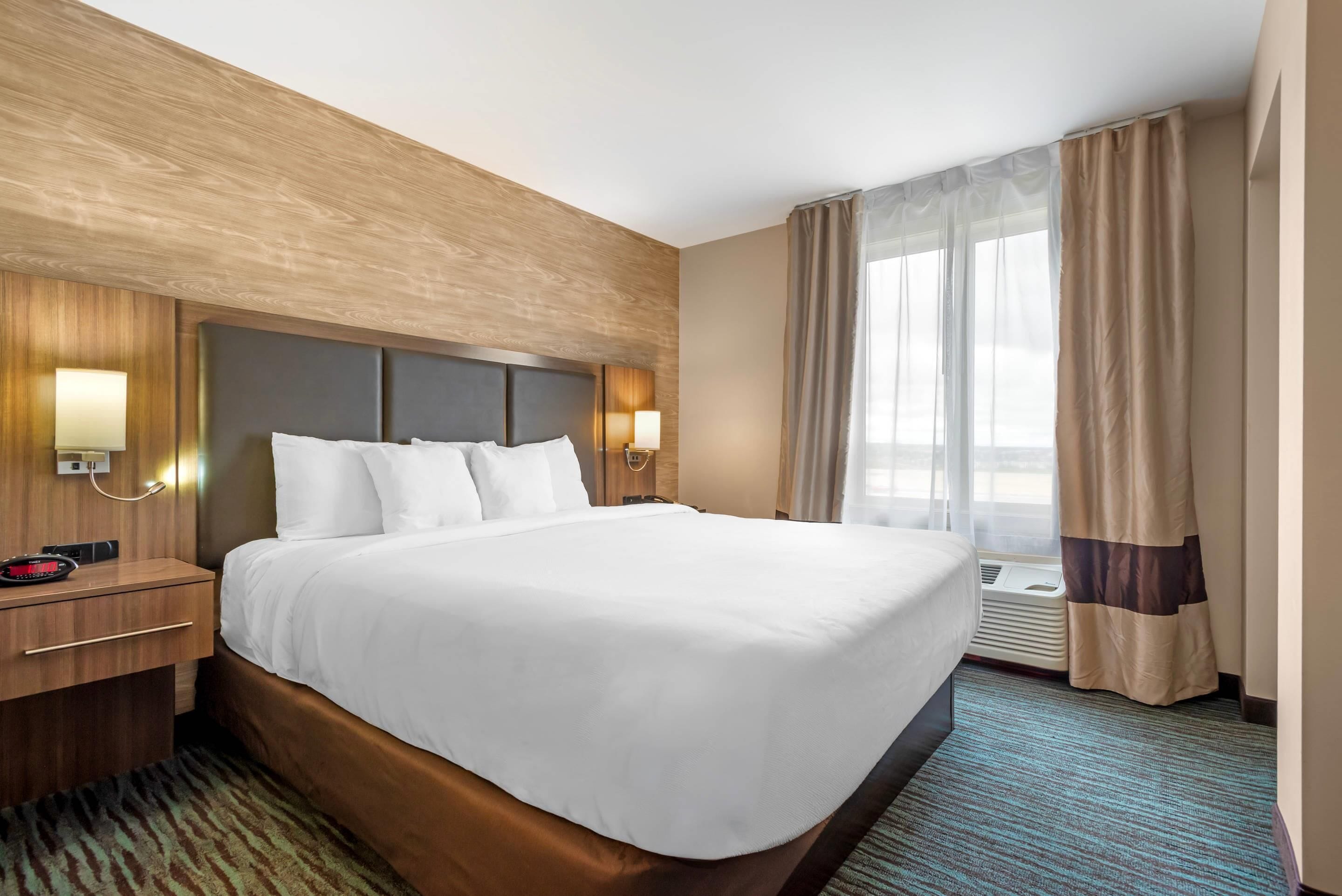 Una habitación de hotel con una cama grande, cabecera de madera, dos lámparas de noche, una mesita de noche y una ventana con cortinas.