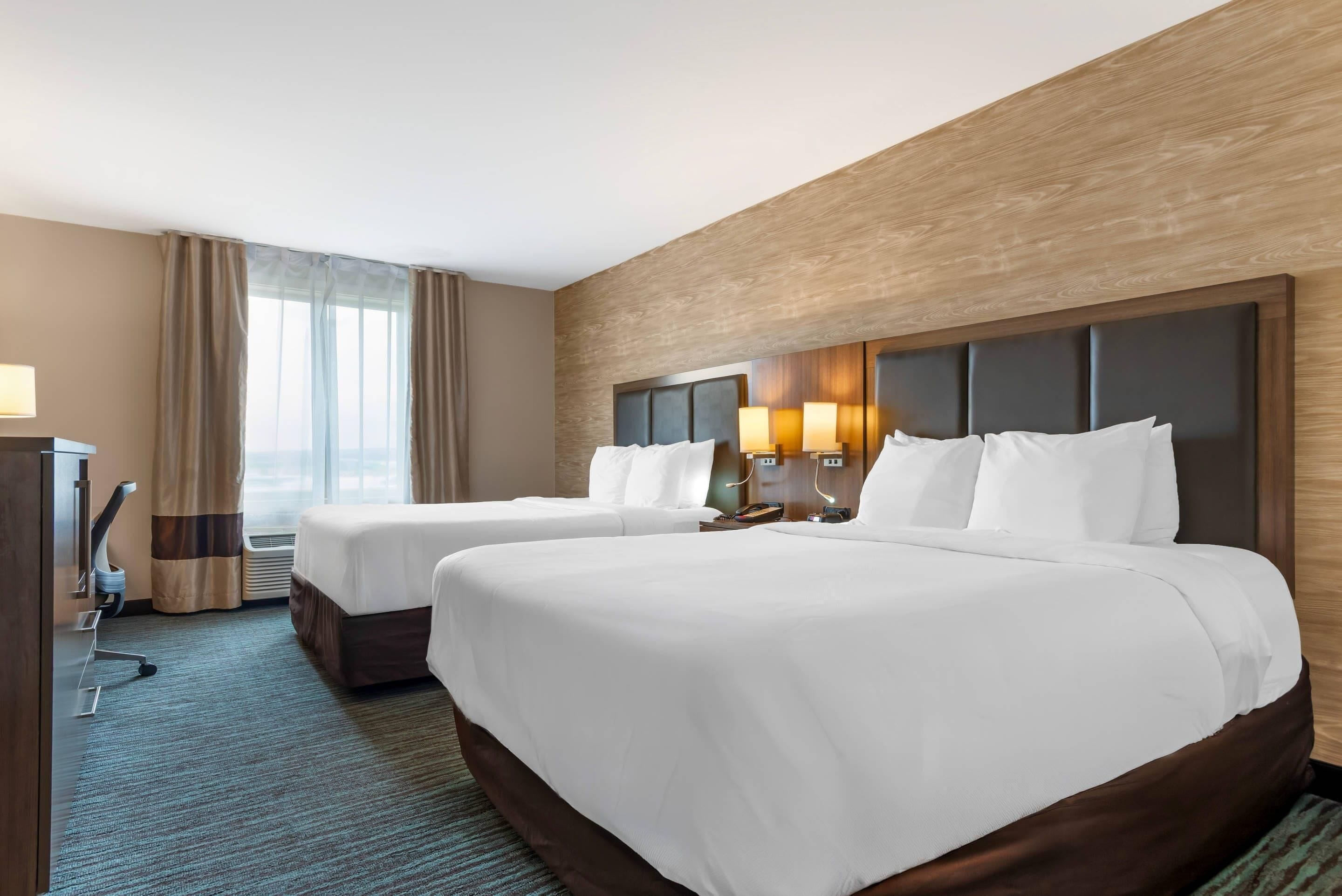 A hotel room with two beds, a desk, a lamp, and a window with curtains.
