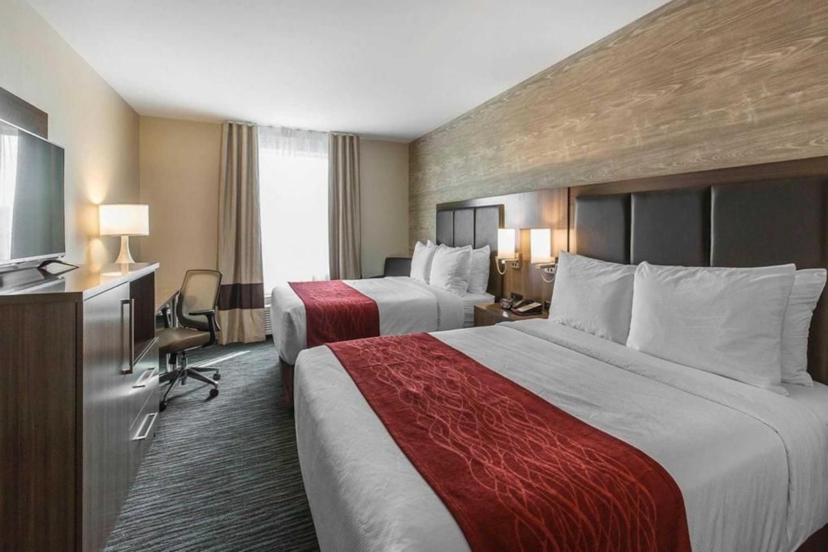 A hotel room with two beds, a desk, a chair, a TV, and a large window with curtains.