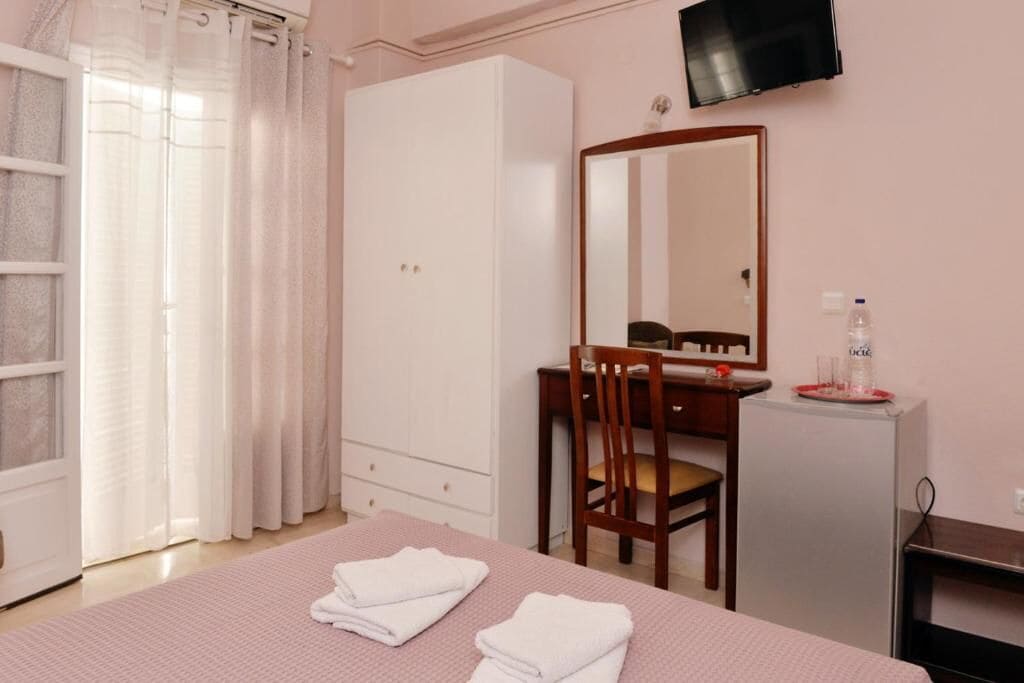 Un dormitorio con cama, escritorio, silla, armario, espejo, televisor y un refrigerador pequeño.