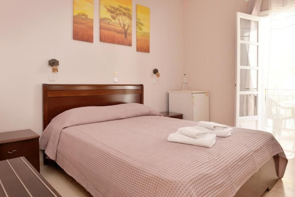 Un lit bien fait, recouvert d’une couvre-lit rose, une table de chevet avec une lampe et une fenêtre donnant sur l’extérieur.