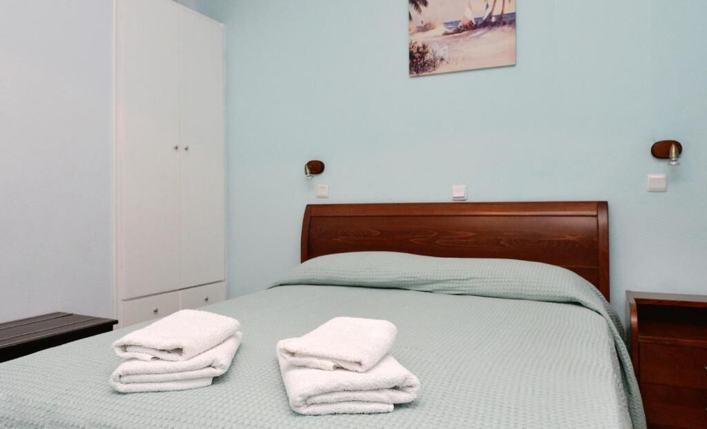 A neatly made bed with a green bedspread and two folded white towels. There is a wooden nightstand with a lamp on it, a white wardrobe, and a painting on the wall.