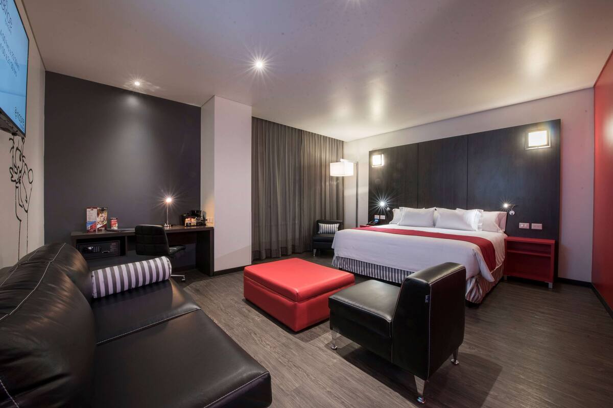 A modern hotel room with a large bed, a red ottoman, a black leather chair, and a desk with a lamp.