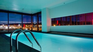 A modern indoor swimming pool with clear glass walls, offering views of a cityscape at dusk. - Ramada Encore by Wyndham San Luis Potosi (San Luis Potosi)