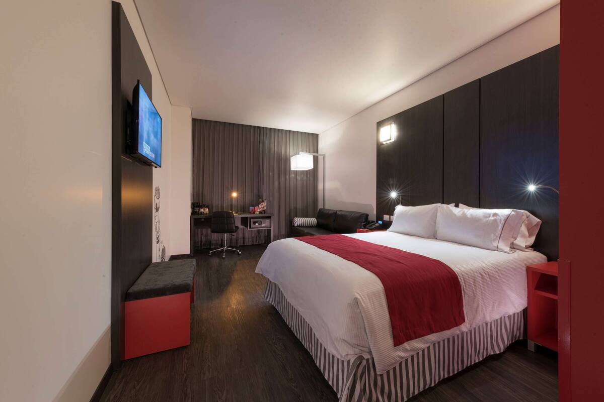 A modern hotel room with a large bed, a desk, a chair, and a TV mounted on the wall.