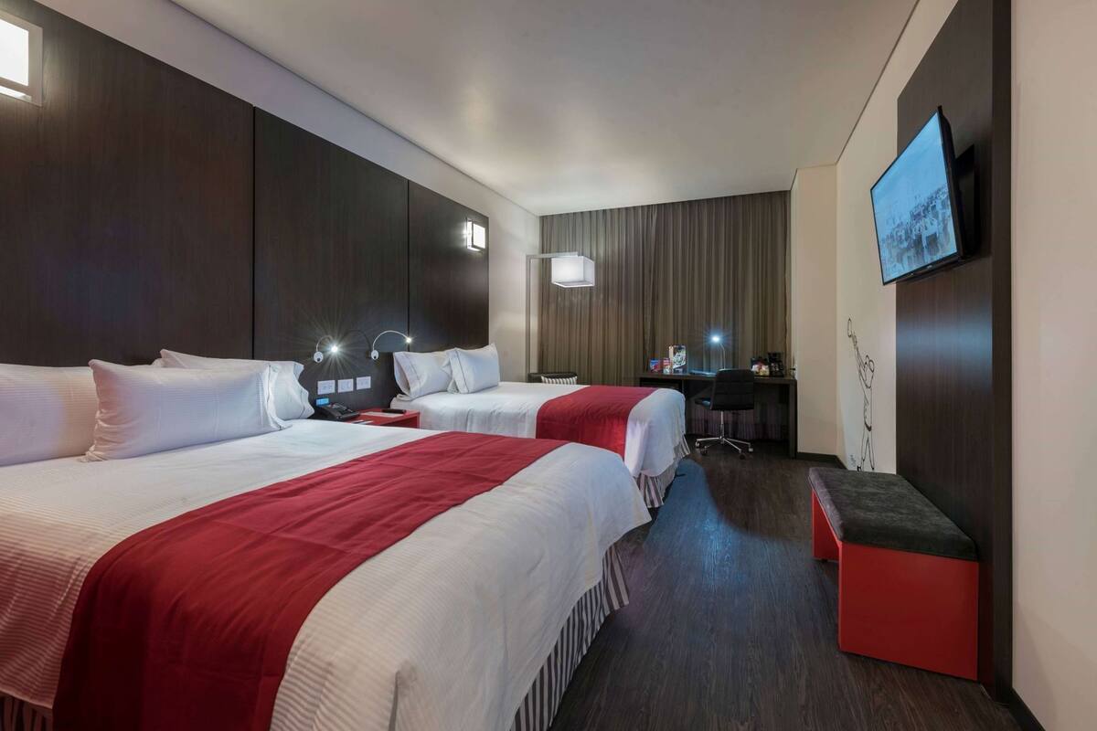 A hotel room with two beds, a desk, a chair, a TV, and a small red ottoman.