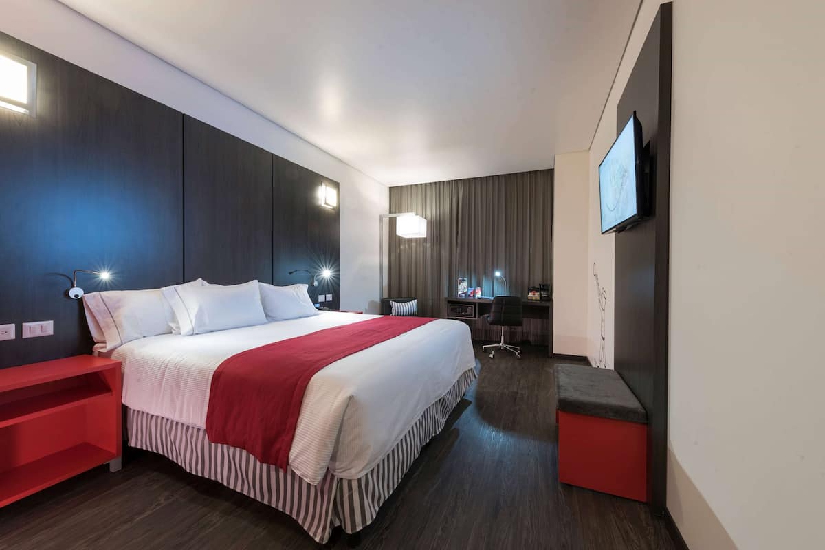 A modern hotel room with a large bed, a desk, a chair, and a TV mounted on the wall.