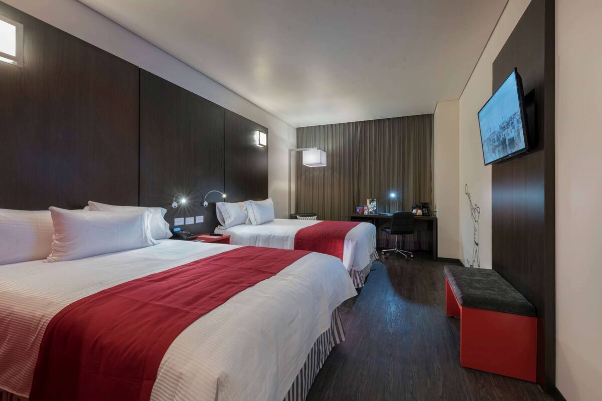 A hotel room with two beds, a desk, a chair, a TV, and a bench.