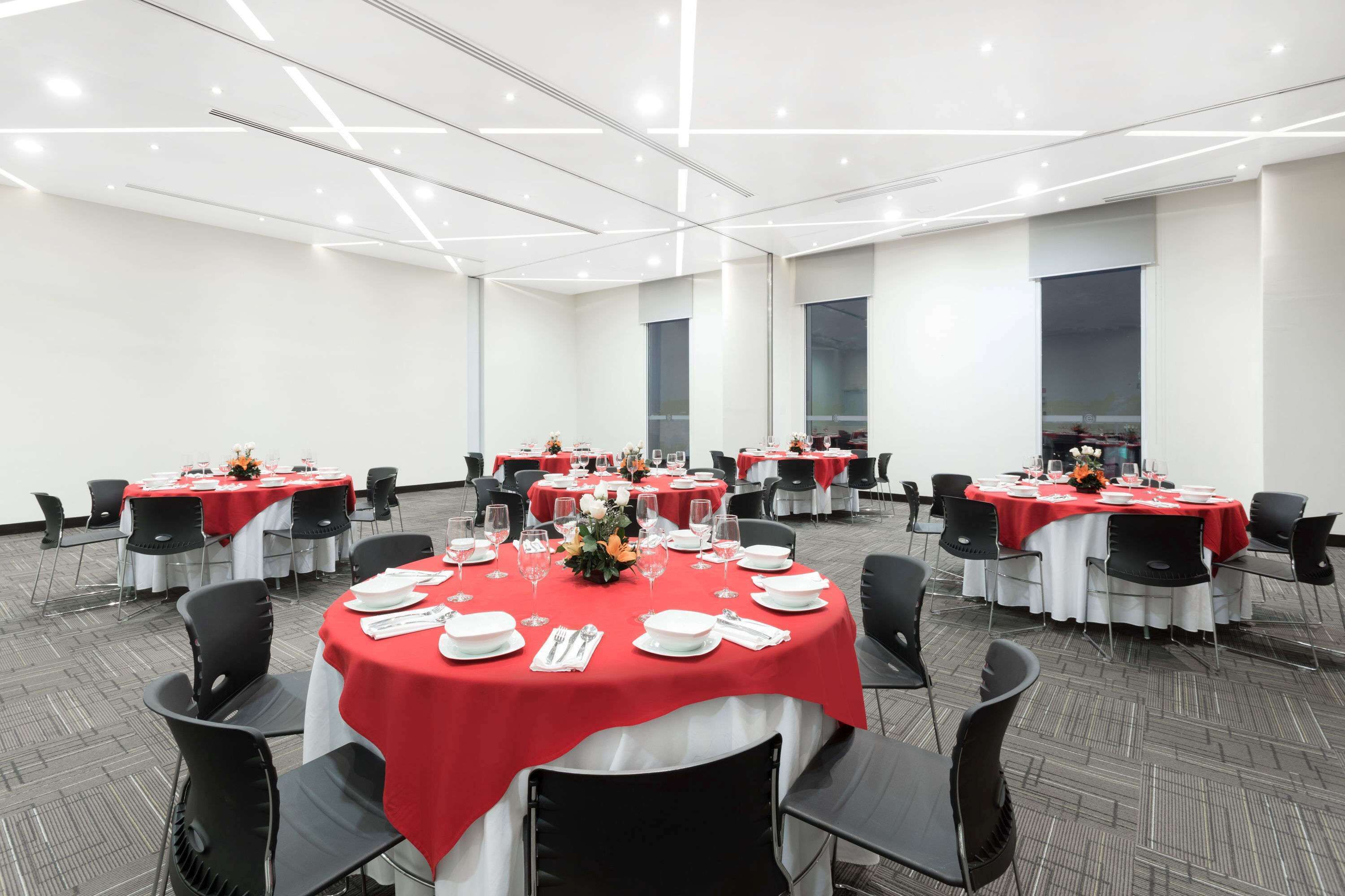A conference room with multiple round tables, each covered with a red tablecloth and surrounded by black chairs.