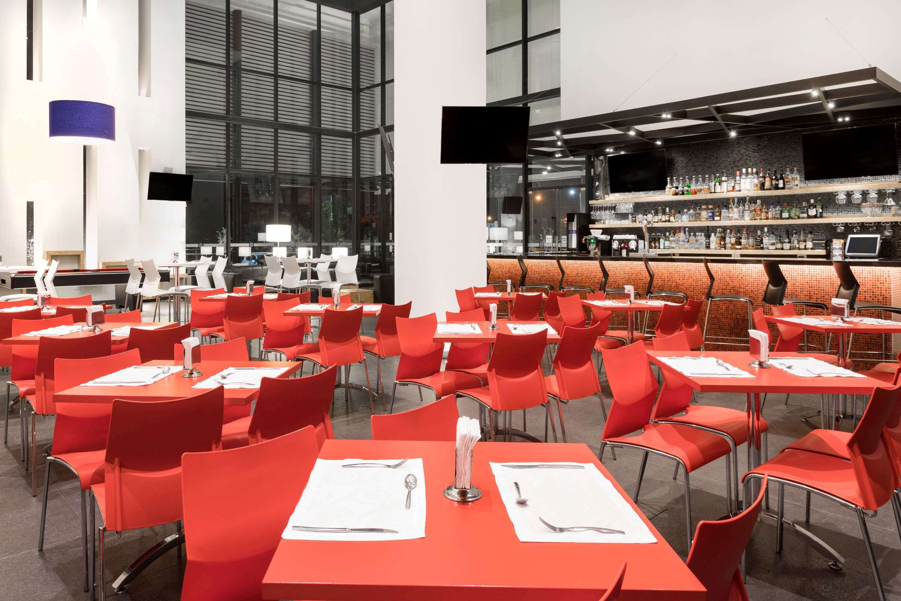 A modern restaurant with red chairs and tables, a bar area with shelves of bottles, and large windows.