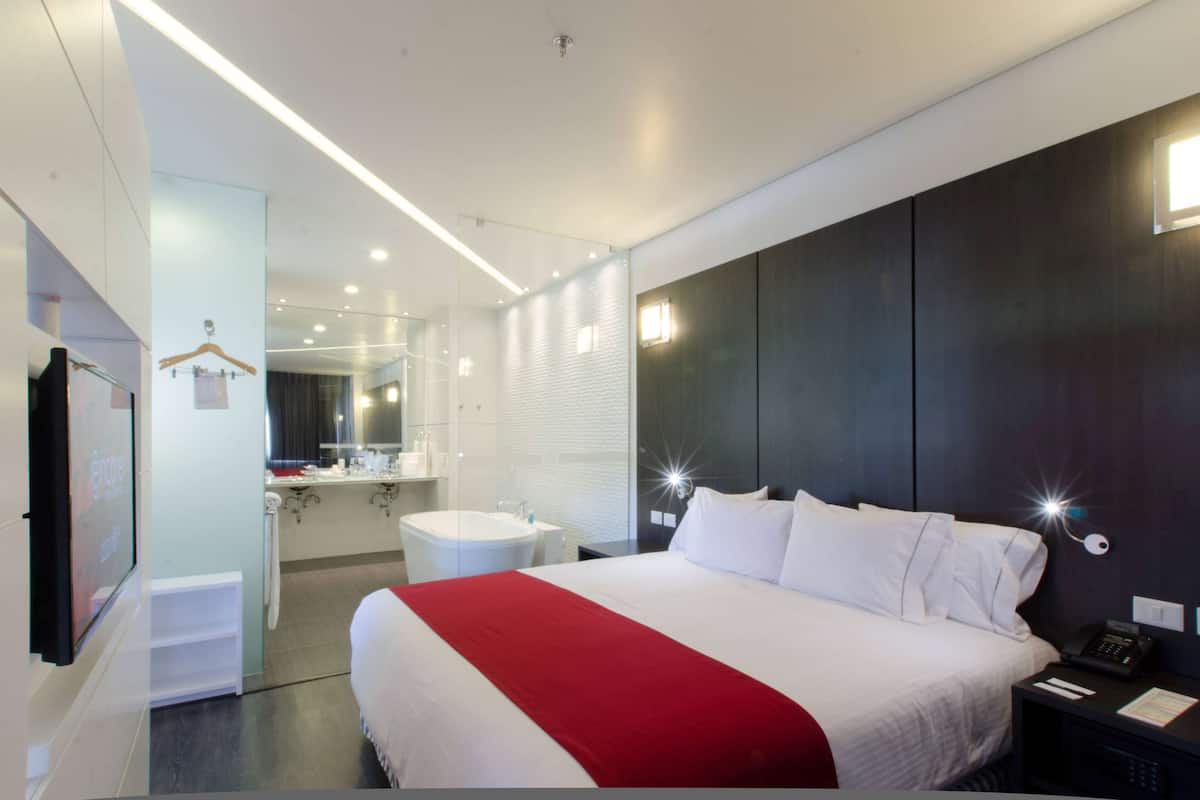 A modern hotel room with a large bed, a bedside table with a phone, a mirror, a bathroom with a bathtub, and a wardrobe.