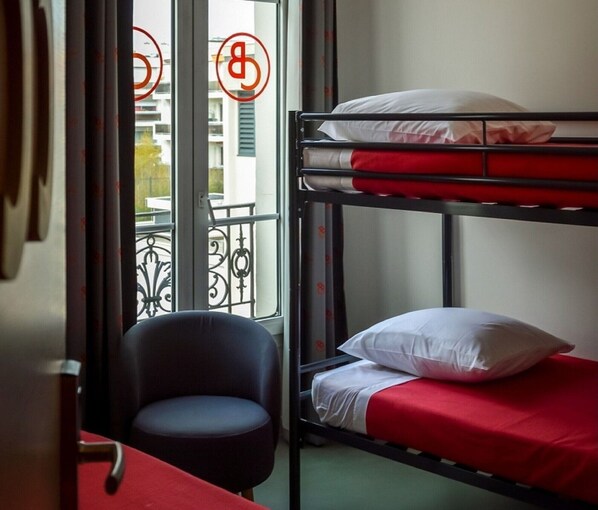 Una cama individual con ropa de cama roja, una silla y una ventana con vistas a edificios.