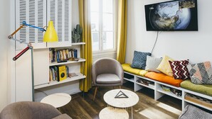 A modern living room with a sofa, armchairs, a bookshelf, a TV, and a window with curtains. - Beautiful Belleville Hostel & Hotel (Paris)