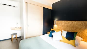 A hotel room with a bed, a small stool, a bedside table, and a wall-mounted air conditioner.