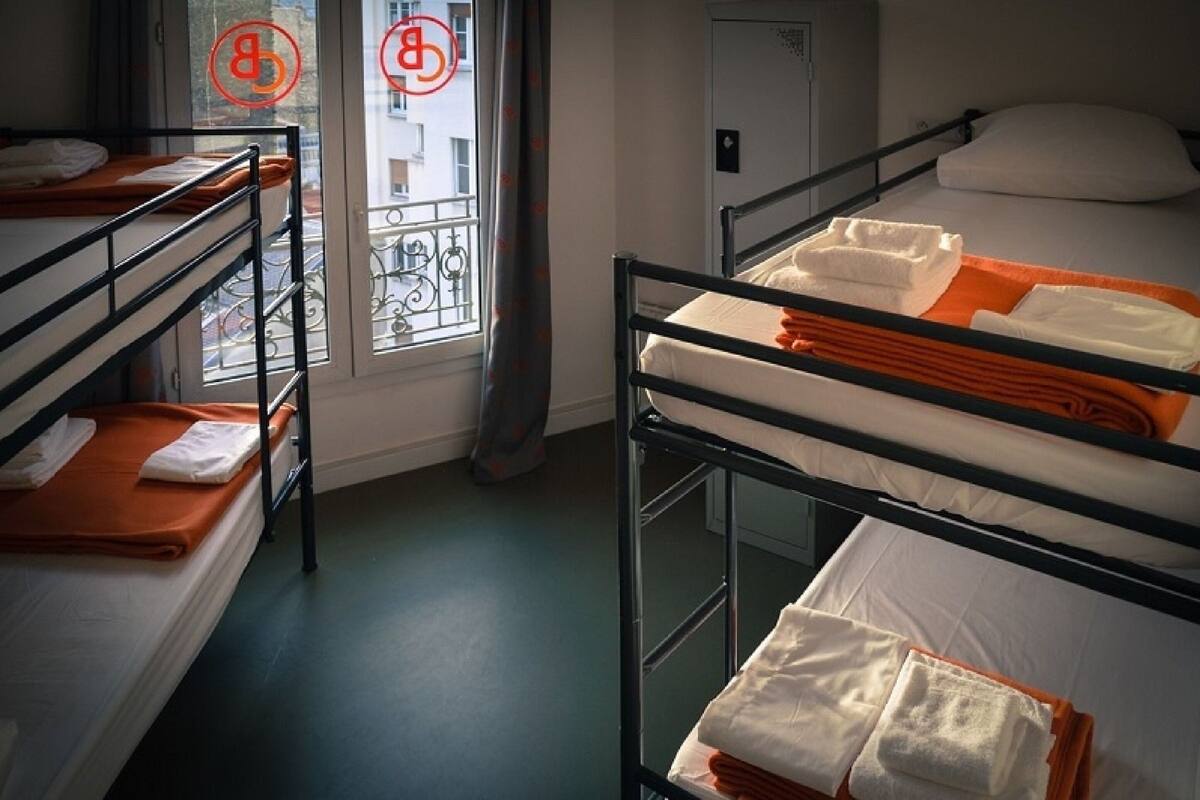 A dormitory room with bunk beds, a window with a view of buildings, and a door.