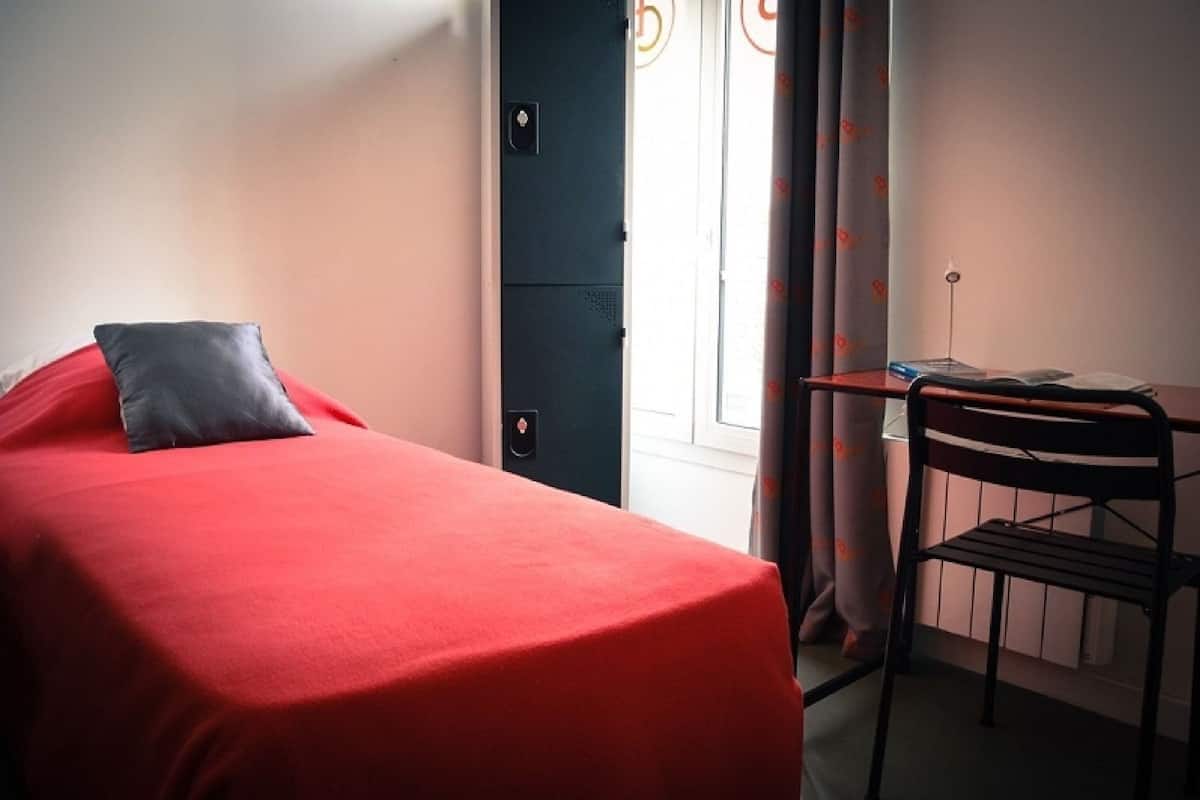 A single bed with a red cover and a gray pillow, a small desk with a chair, and a door with a handle.