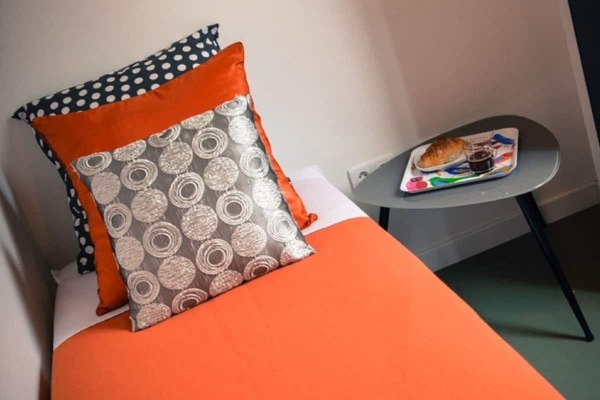 A bed with orange bedding and patterned pillows, a small table with a tray of food, and a grey wall.