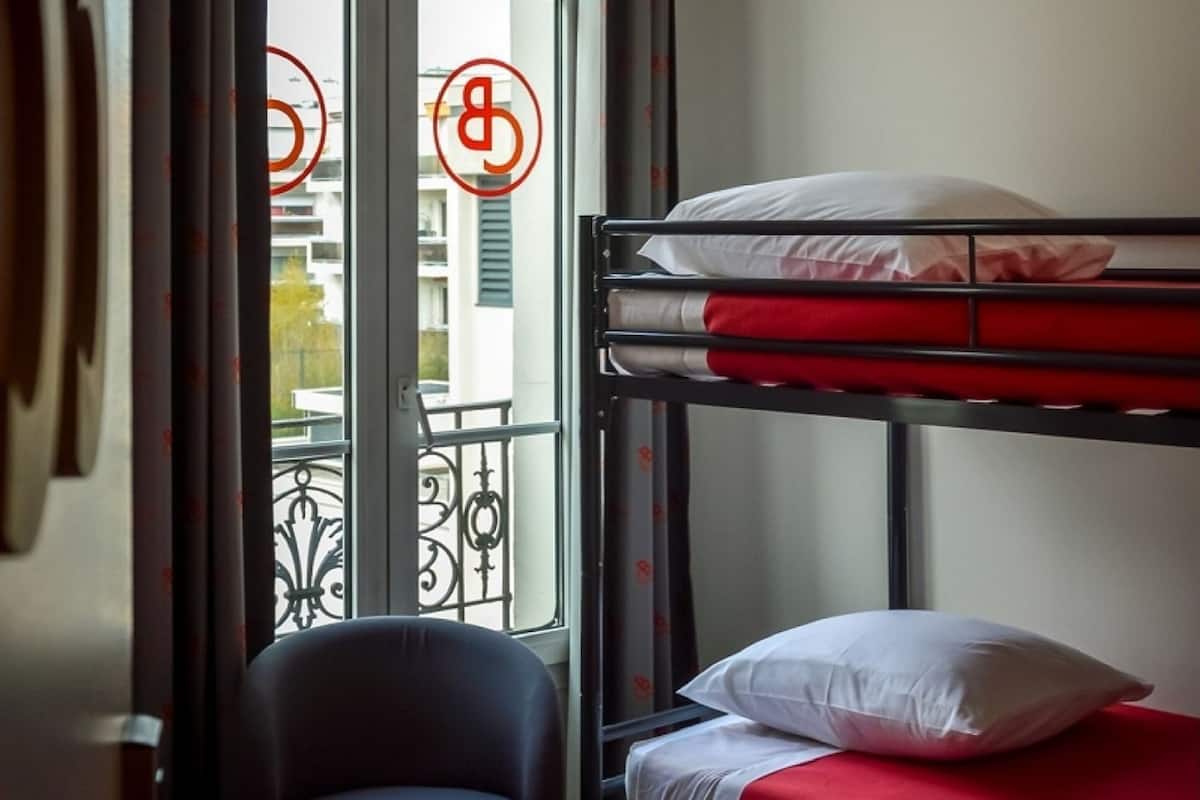 A bunk bed with red bedding and a chair in a room with a view of buildings through a window.