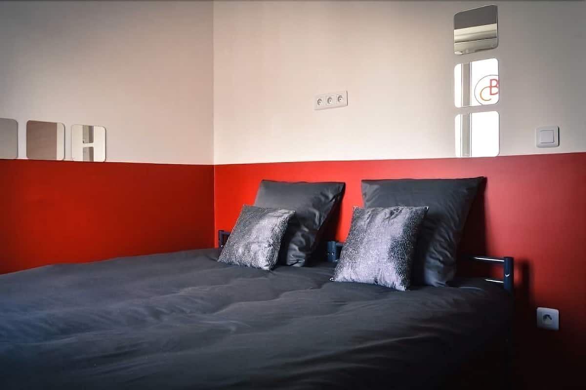 A room with a bed against a red wall, a window with blinds, and a thermostat.