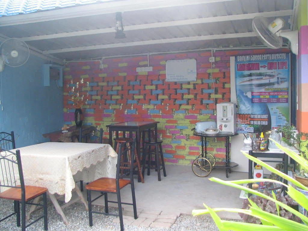 Uma pequena área externa coberta com mesa e cadeiras, uma parede de tijolos colorida e um ventilador.